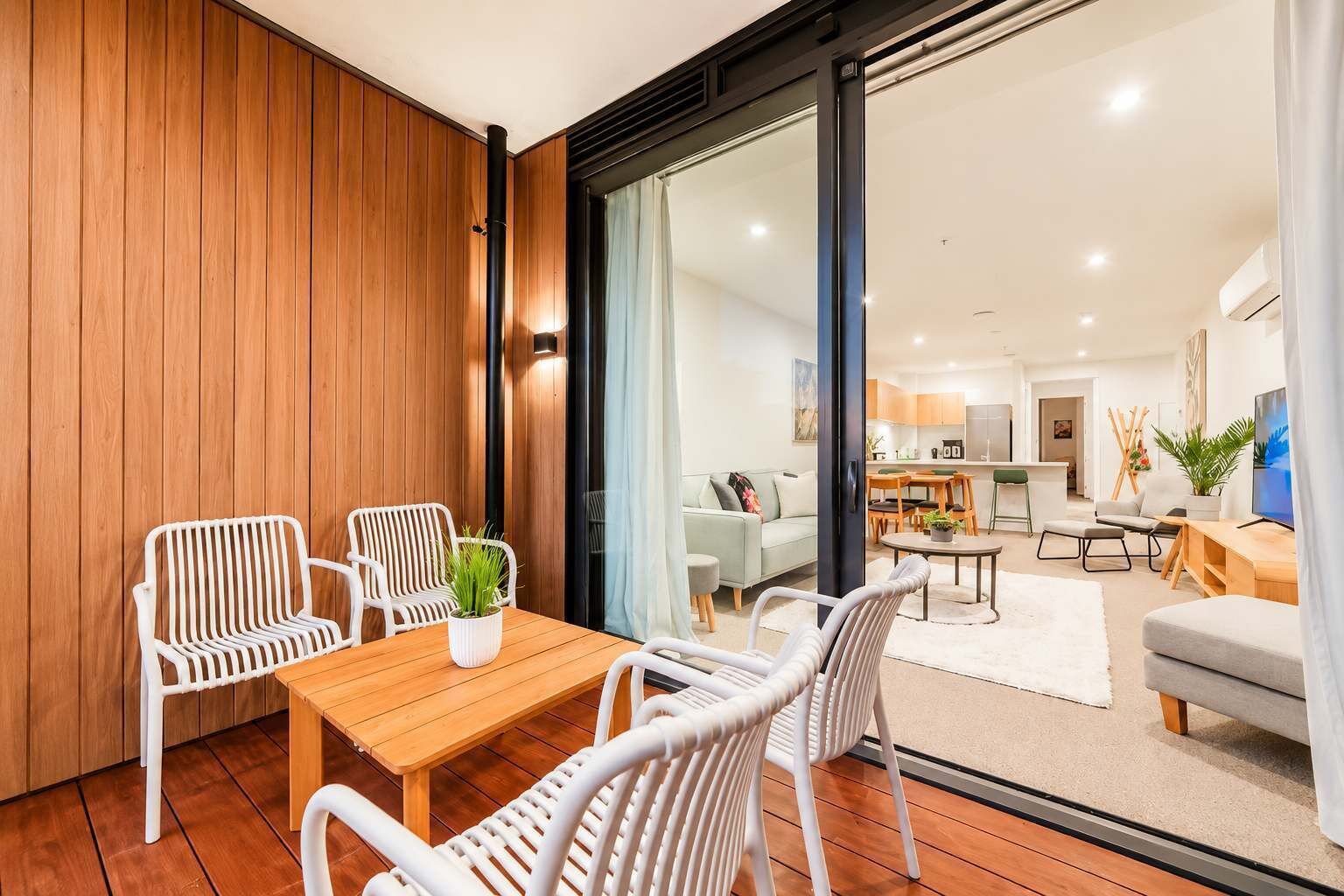 A modern living room with a wooden floor, white wicker furniture, and a view into the kitchen area.