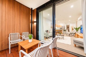 A modern living room with a wooden floor, white wicker furniture, and a view into the kitchen area.