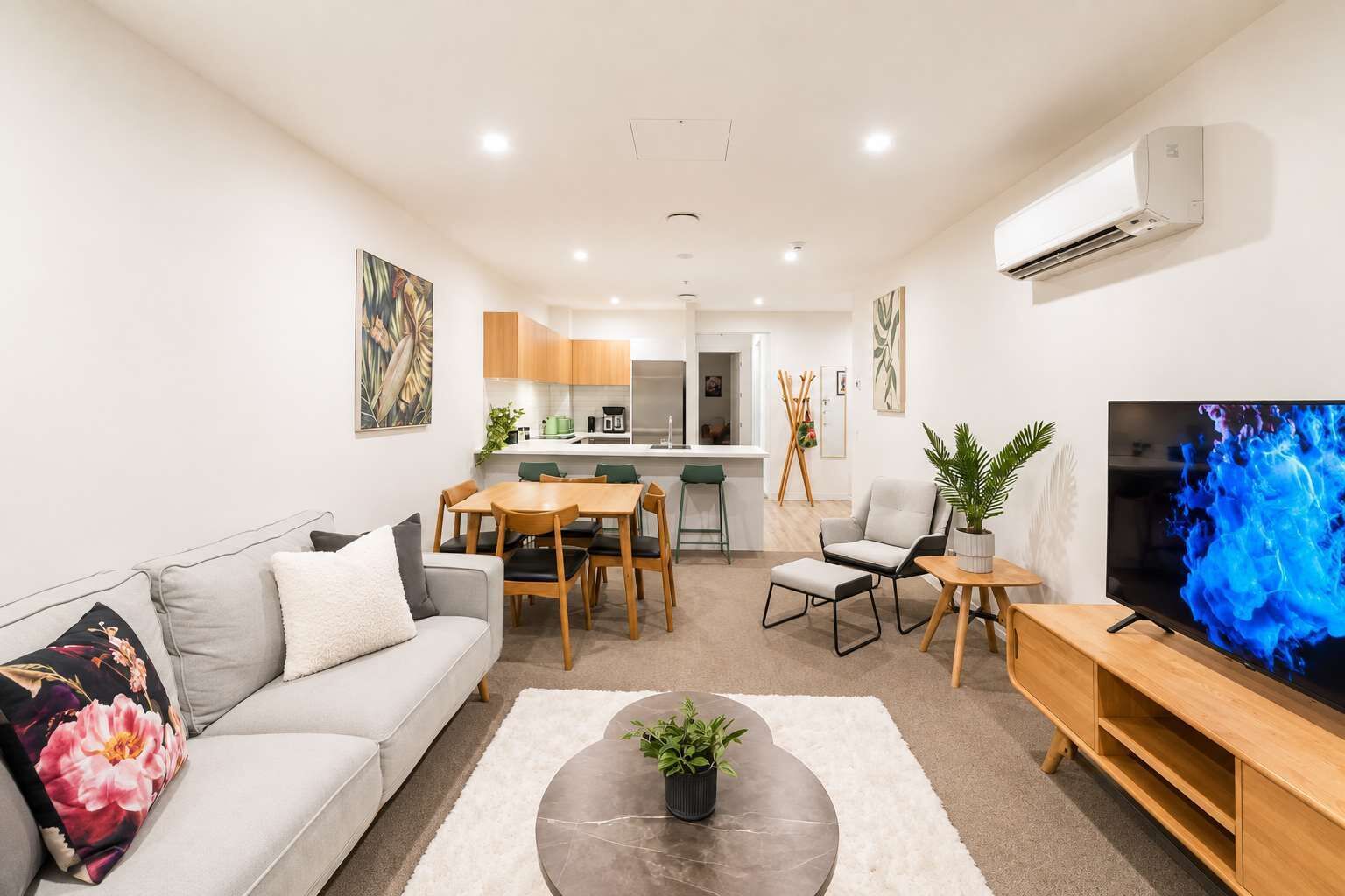 A modern living room with a sofa, coffee table, and TV. There is a dining area with a table and chairs, and a kitchen in the background.