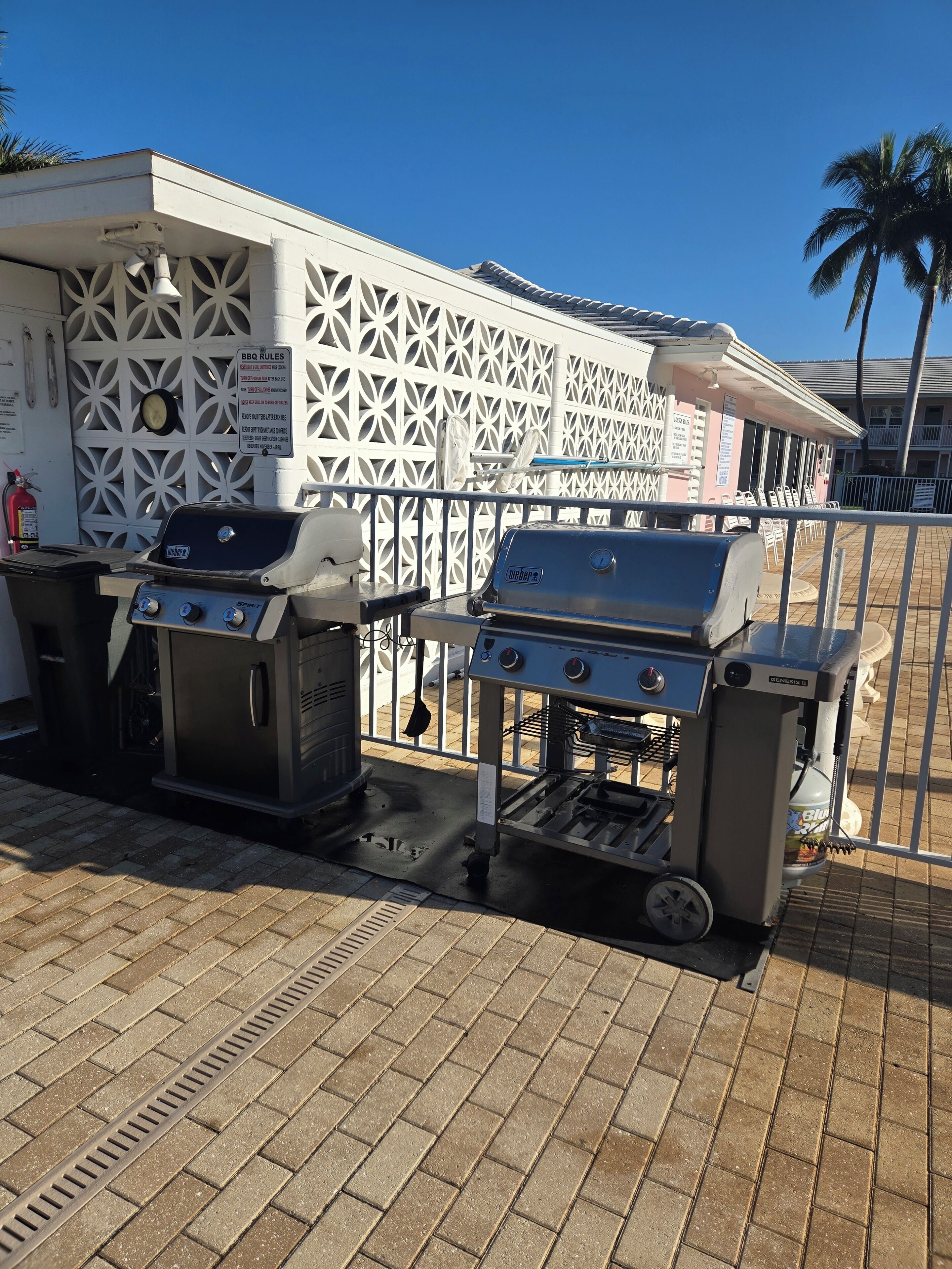 Una zona de barbacoa con dos parrillas, un edificio blanco con fachada decorativa y una palmera al fondo.