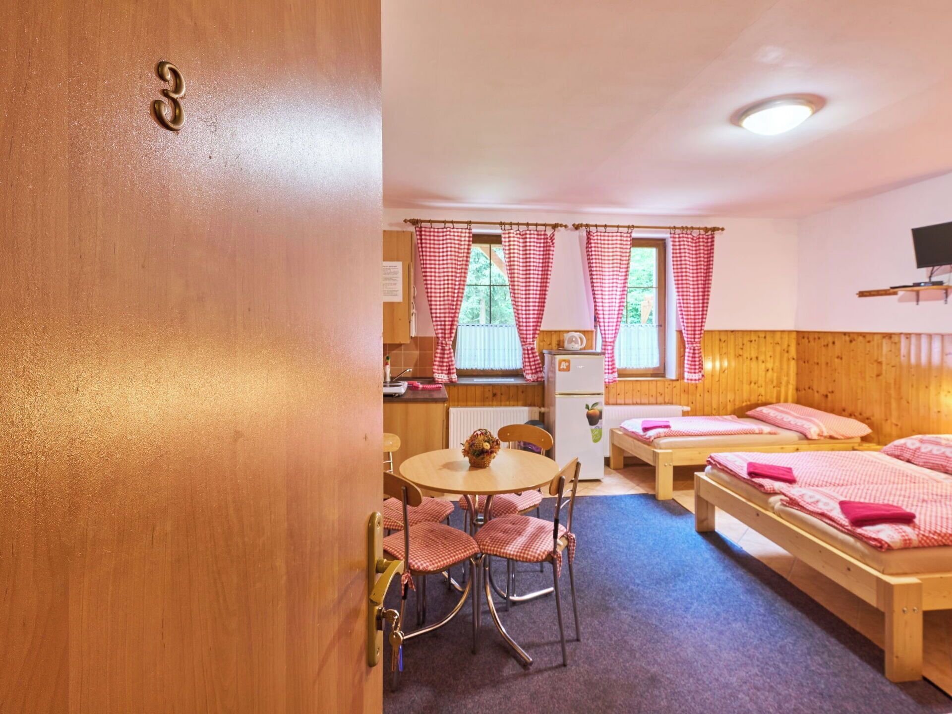 Habitación con dos camas de madera, una mesita con sillas, una zona de cocina y una ventana con cortinas a cuadros rojos y blancos.