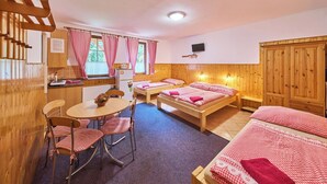 A cozy room with two wooden beds, a small dining table with checkered chairs, a kitchen area with a refrigerator, and a TV mounted on the wall.