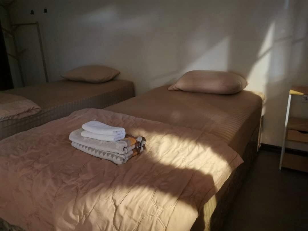 Une chambre d’hôtel avec deux lits, chacun recouvert d’une couverture et doté d’un oreiller de couleur beige. On y trouve une petite table avec une lampe et une chaise.