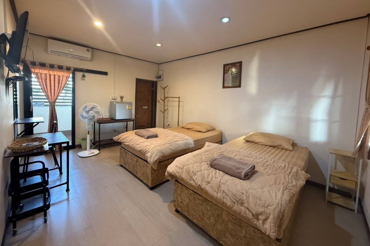 A room with two beds, a desk, a chair, a fan, and a window with curtains.