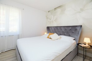 Una cama bien tendida con sábanas blancas y cabecera gris, una mesita de noche con lámpara, y una ventana con cortinas traslúcidas.