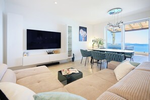 Una sala de estar moderna con un televisor de pantalla plana grande, asientos cómodos, un comedor con sillas y vistas al mar a través de amplios ventanales.