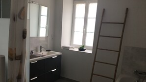 Een badkamer met een witte wasbak, donkere kastjes, een spiegel, een raam met een plant en een houten ladder.