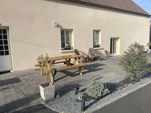 Ein Gebäude mit sandfarbener Fassade, eine gepflasterte Terrasse mit einem Holztisch und Stühlen sowie Topfpflanzen.