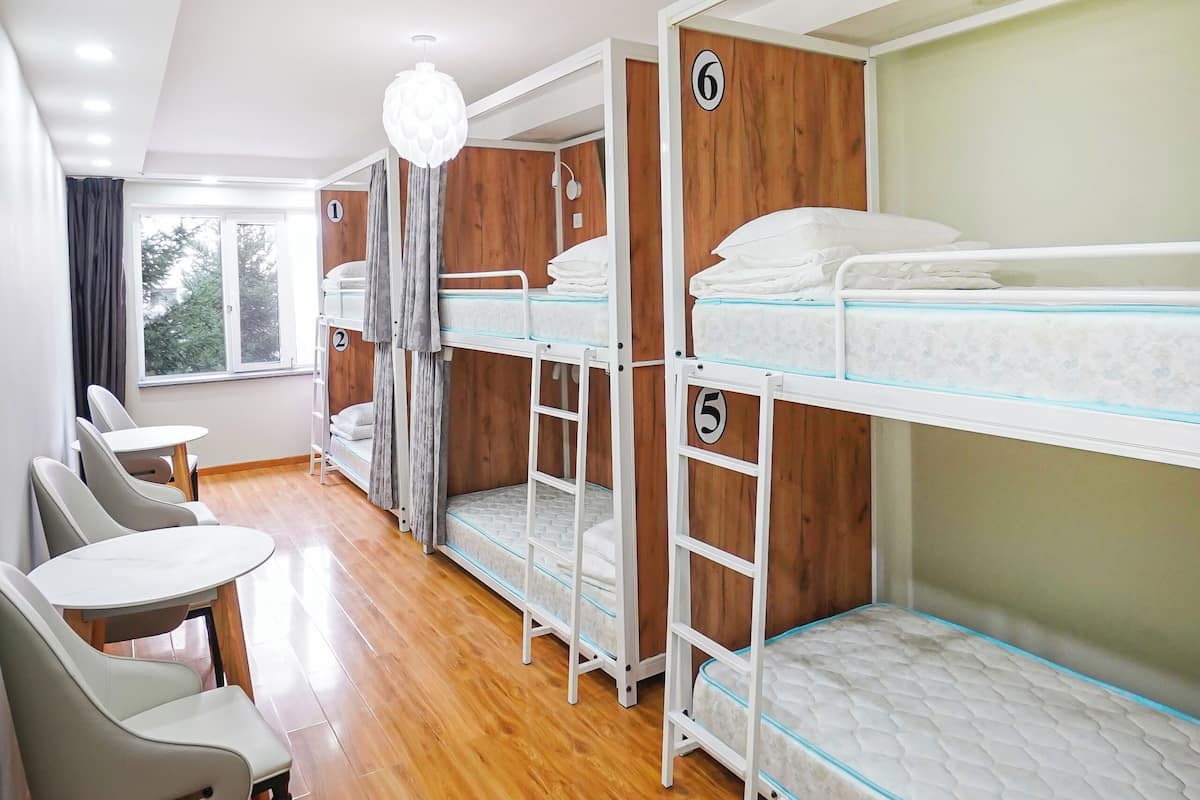 A dormitory room with bunk beds, a wooden floor, a table with chairs, and a window with curtains.