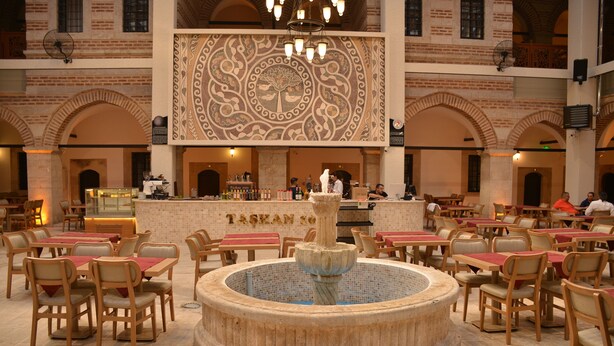 Un vaste espace de restauration intérieur, doté de nombreuses tables et chaises, d’une fontaine centrale et d’un mur décoratif orné de motifs complexes.