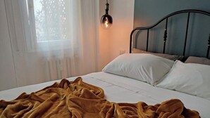 Una cama con colchón y almohadas blancos, una colcha color mostaza y un cabecero de metal negro, además de una lámpara colgante.
