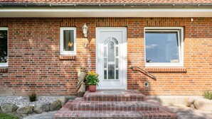 Une maison en briques avec une porte blanche, deux fenêtres, un petit porche avec des marches et un pot de fleurs.