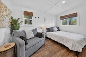 Ein Zimmer mit grauem Sofa, einem weißen Bett, Holzboden und einem Fenster mit Blick auf Bäume.