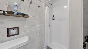 Un baño con ducha, inodoro y lavabo. Se aprecia un perchero con toalla y una botella de jabón de manos.