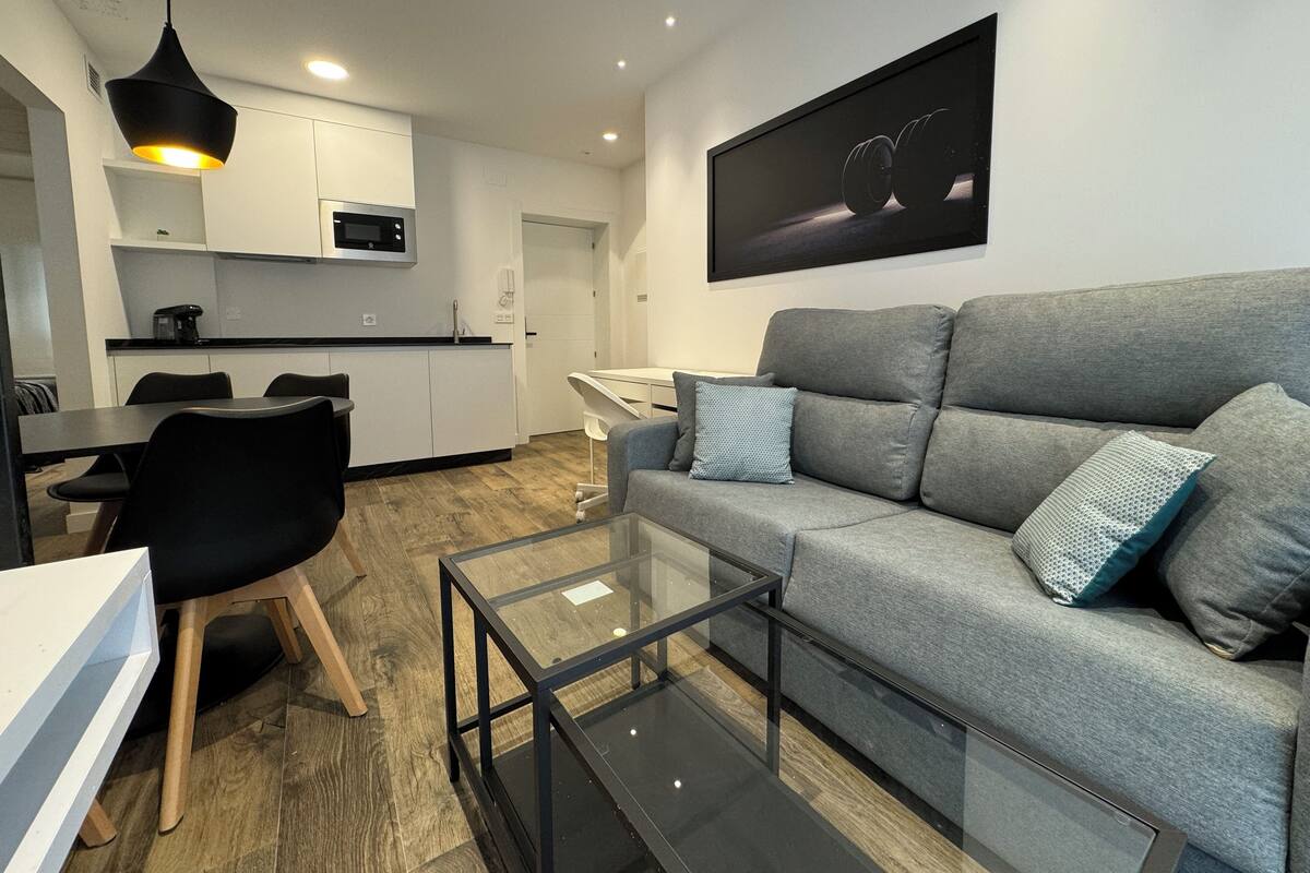 Ein modernes Wohnzimmer mit einem grauen Sofa, einem Glastisch und einem Essbereich mit einem weißen Tisch und schwarzen Stühlen.