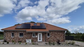 Uma casa térrea de tijolos com telhado de cerâmica, painéis solares e um pequeno jardim.