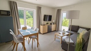 Uma sala moderna com sofá, mesa de jantar com cadeiras, TV fixada na parede e área de cozinha com micro-ondas.