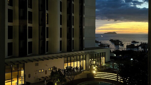 Ein Hochhaus-Hotel mit Poolbereich auf dem Dach, großen Fenstern und Blick auf das Meer bei Sonnenuntergang.