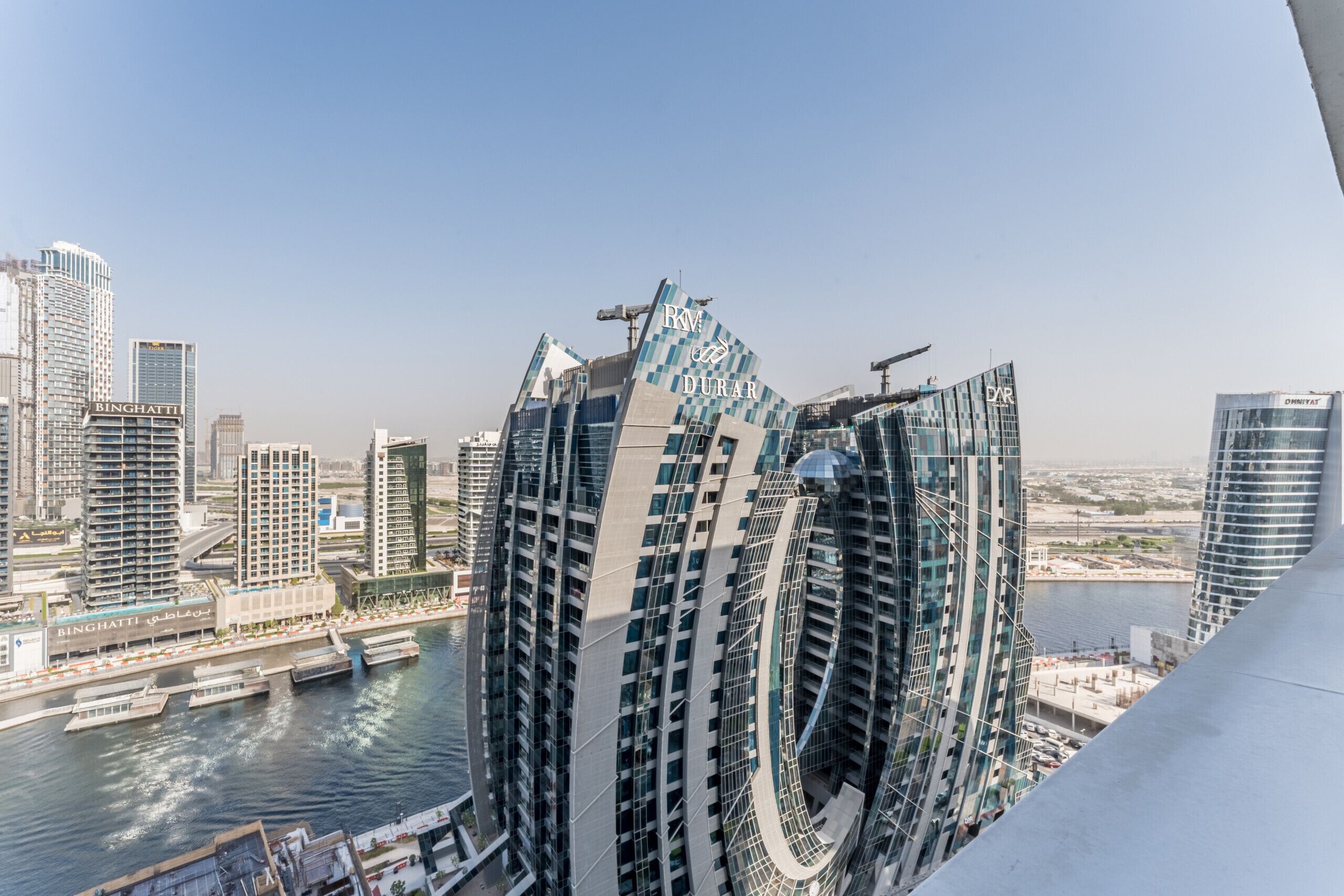 A modern skyscraper with a curved facade and a crane on top, overlooking a body of water and other buildings.