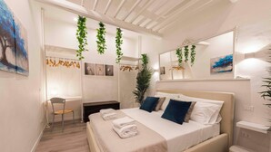 A modern bedroom with a large bed, a desk, and a chair. There are plants and artwork on the walls. - Deluxe Pitti Suites by MMega (Florence)