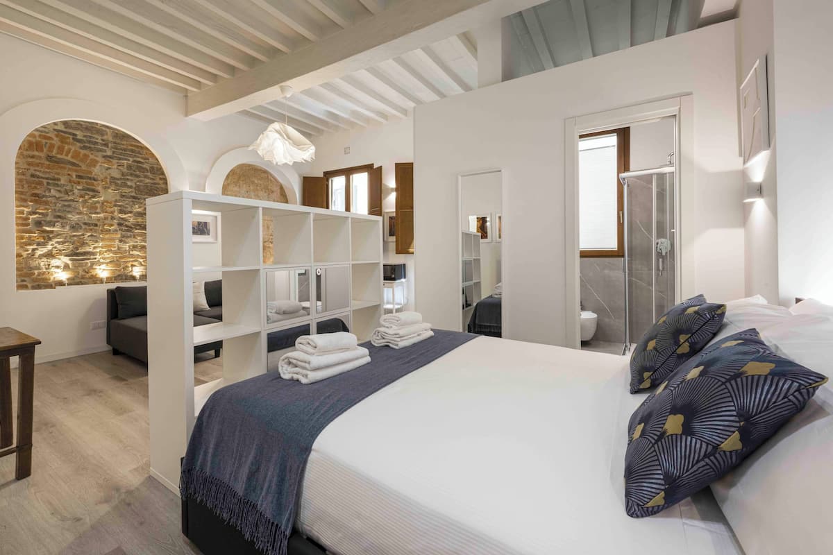 A modern bedroom with a large bed, built-in shelving, a stone wall, and a shower area.