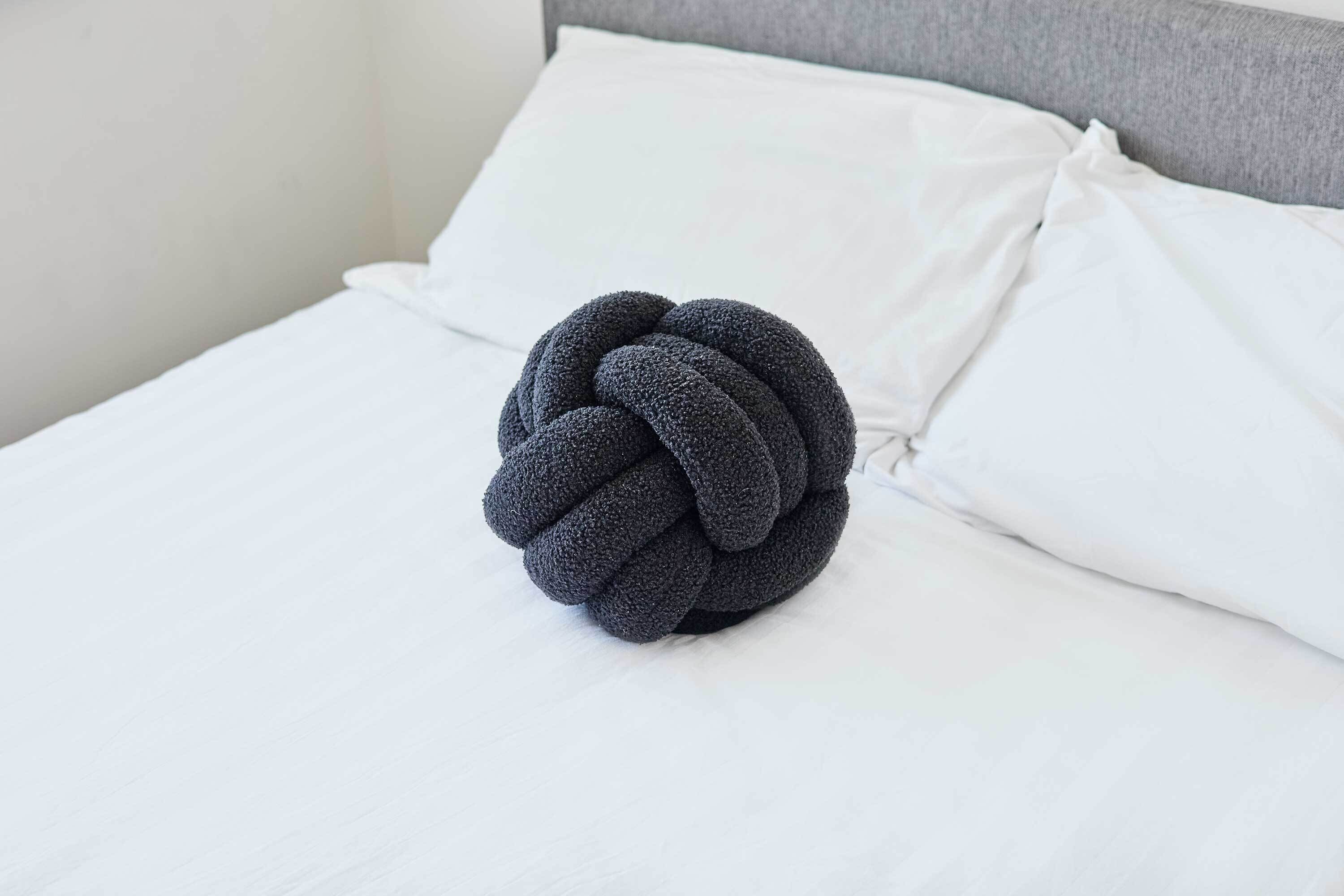 Ein Bett mit weißer Bettwäsche und einem dunkel, strukturierten Kissen in knopfartiger Form.