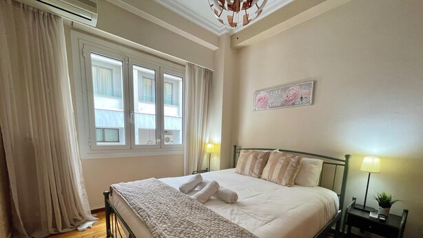 Een net opgemaakt bed met een sprei, twee bedlampen, een wanddecoratie en een raam met gordijnen.