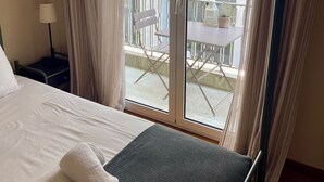 Een hotelkamer met een bed, nachtkastje, lamp en uitzicht op een balkon met tafel en stoelen.