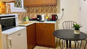Een praktische keuken met een magnetron, kookplaat, houten keukenkastjes, een betegelde spatwand en een kleine eettafel met een stoel.