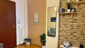 Een kamer met een houten vloer, een grijze bank, een witte tafel, een spiegel, een radiator, een deur, een muur met een inspirerende quote, en een plank met boeken en planten.