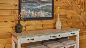 A wooden interior with a white desk and bench, a framed landscape painting, and a vase with flowers. - Budget-Friendly! Creekside, Hot Tub, Firepit (Gatlinburg)