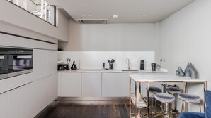 Una cocina moderna con horno empotrado, cafetera y lavaplatos. Hay una zona de comedor con mesa y taburetes de bar.