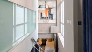 Un pasillo estrecho con un separador de vidrio en un lado, una cama con cabecera y almohadas en el otro, y un cuadro abstracto de gran tamaño en la pared.