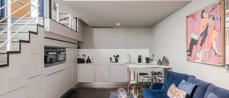 A modern kitchen with built-in appliances, a dining area with a table and chairs, a blue sofa, and a staircase.