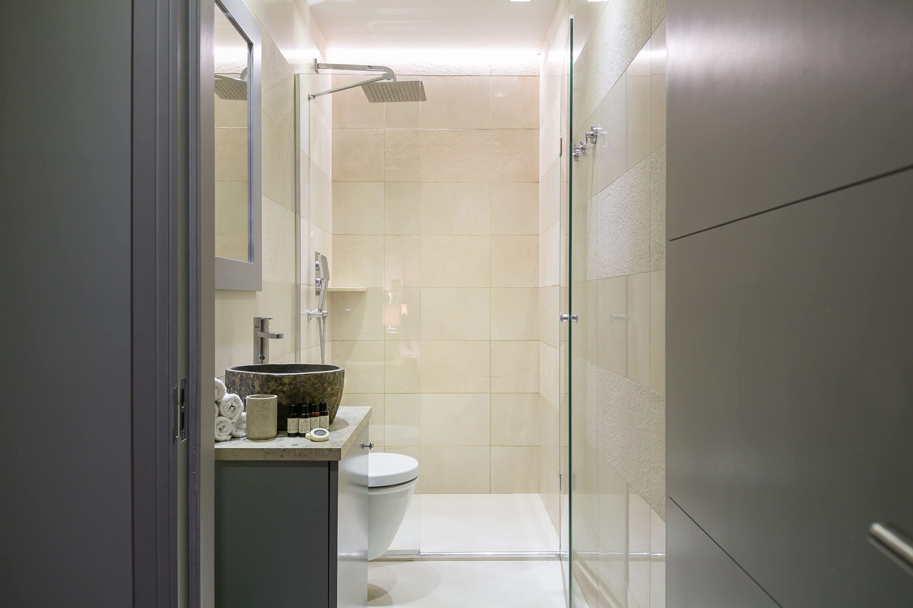 Moderni kylpyhuone, jossa on lasinen suihkuseinä, wc ja pesuallas, jonka päällä on hygieniatuotteita.