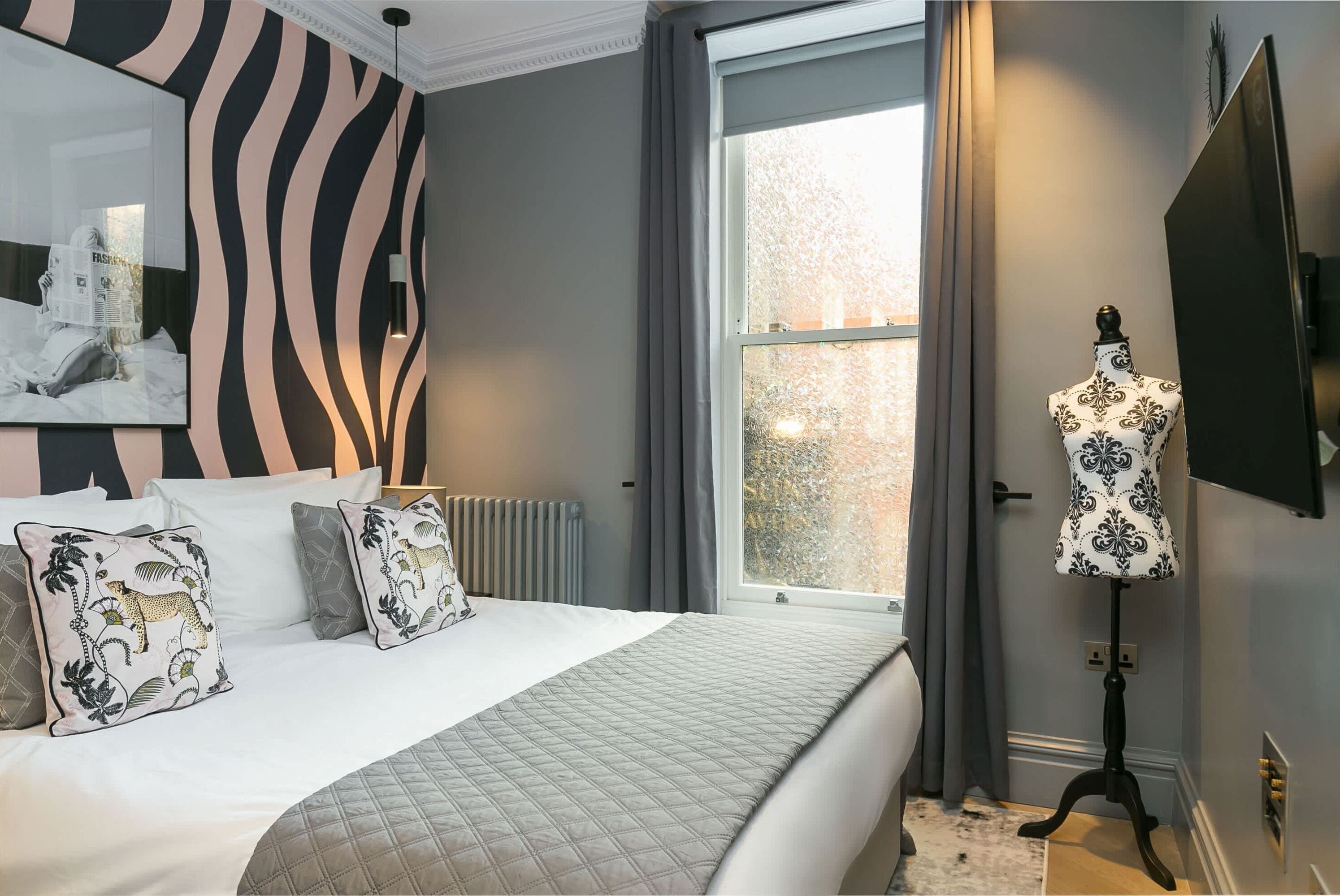 Une chambre avec un grand lit, un mur orné d’un motif zébré, une fenêtre avec des rideaux, un lampadaire et un mannequin.