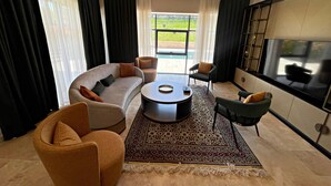 Een moderne woonkamer met een grote bank, een salontafel en twee fauteuils. Er is een groot raam met gordijnen, een kroonluchter en een flatscreen televisie aan de muur bevestigd.