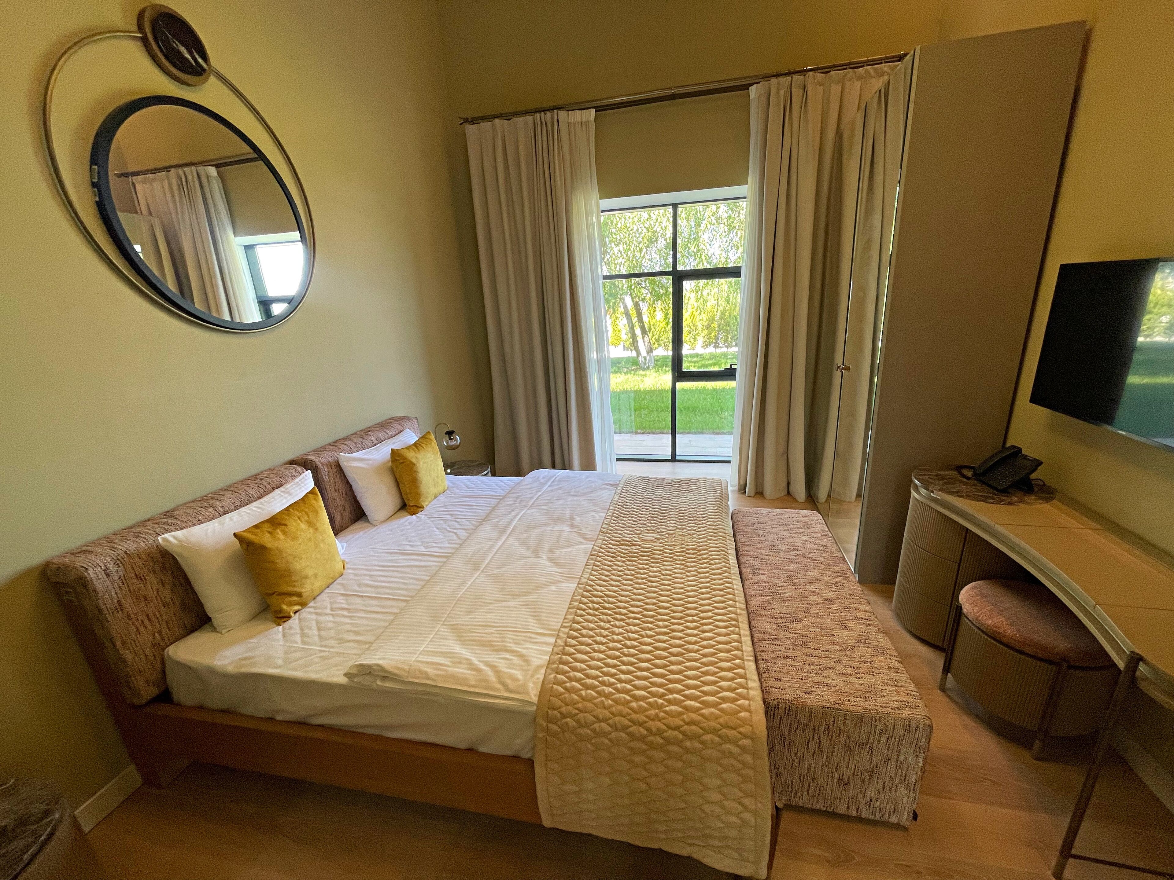 A hotel room with a bed, a desk with a telephone, a television, and a view of a green landscape through the window.