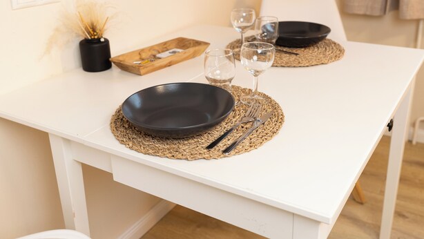 A white dining table with black plates, cutlery, and wine glasses set on woven placemats.