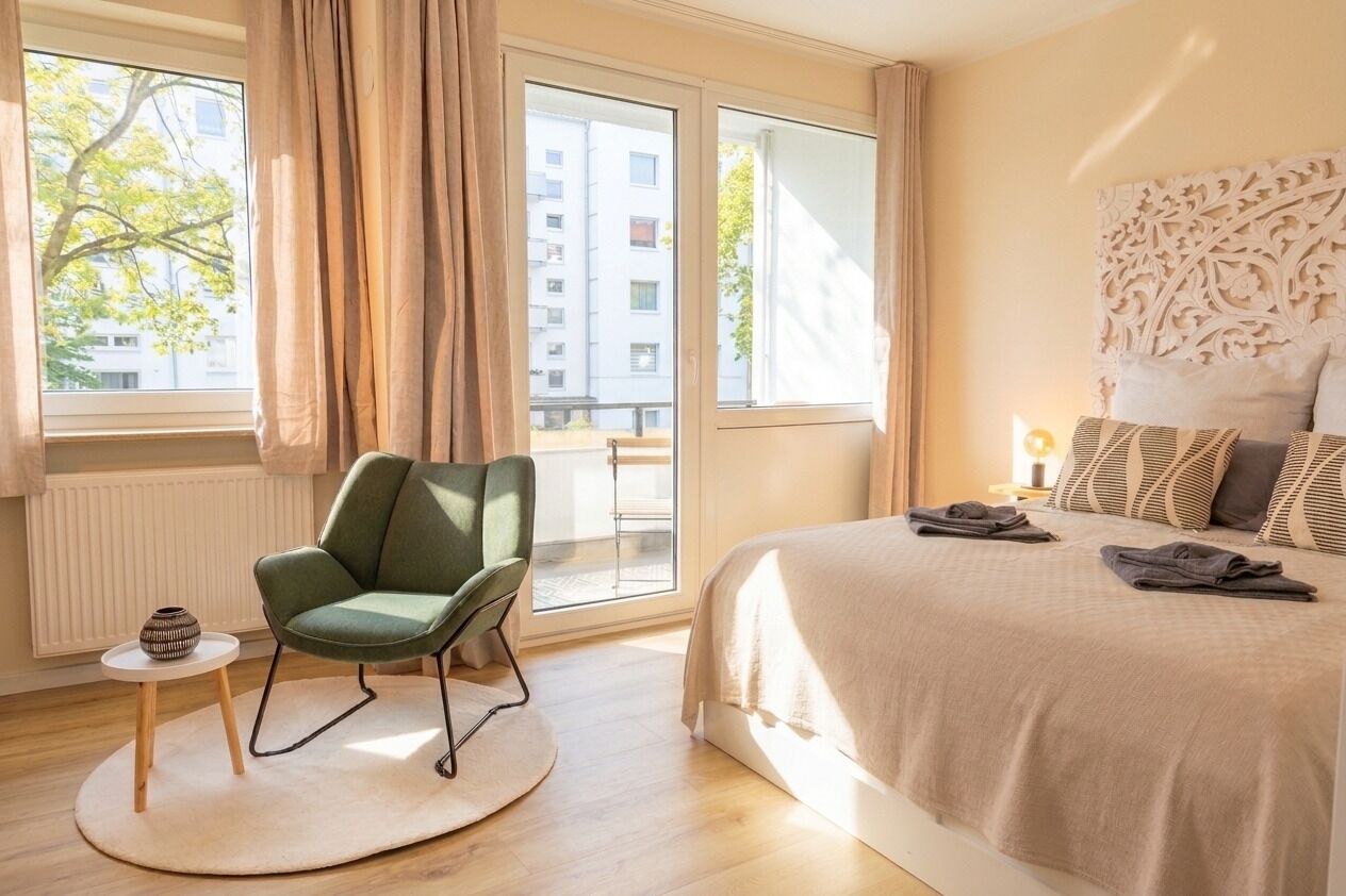 Ein modernes Schlafzimmer mit einem großen Bett, einem grünen Sessel, einem kleinen Tisch und Blick ins Freie.