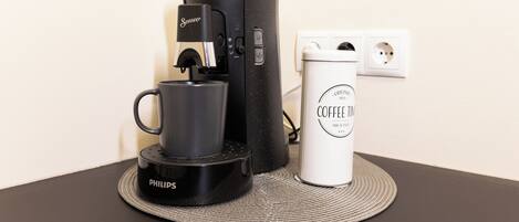 A coffee maker with a cup on a coaster, a white mug, and a power outlet on a table.
