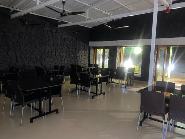 A spacious indoor area with multiple tables and chairs, stone walls, and large windows allowing natural light. - The Paradise Retreat (Akola)