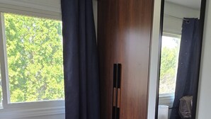 Una habitación con un armario de madera, un espejo, una mesita de noche con lámpara y una ventana con cortinas.