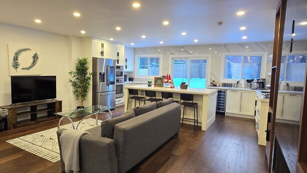Una cocina moderna con una isla central, un área de comedor con barra, y una sala de estar con sofá y televisor.