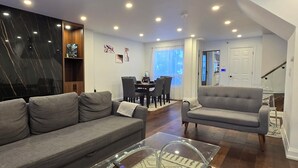 Una sala moderna con un sofá gris, un sillón gris y una mesa de centro de vidrio. Hay un comedor con una mesa y sillas, y un aparador de madera con estantes.