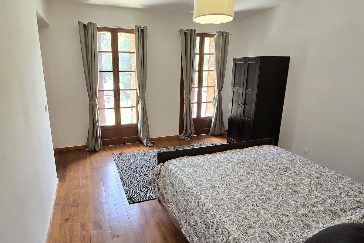 A bedroom with a bed, a wardrobe, a window with curtains, and a wooden floor.