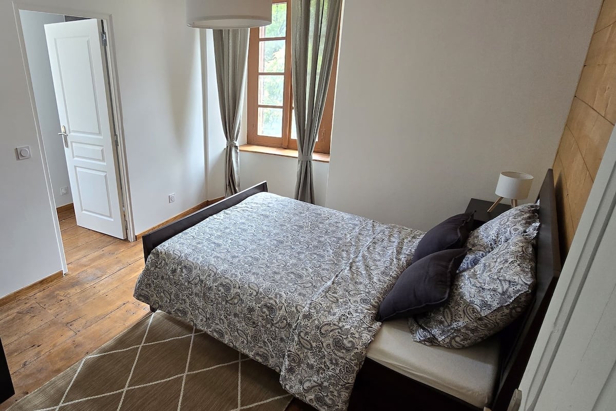 A bedroom with a bed, a window with curtains, a door, and a wooden floor.