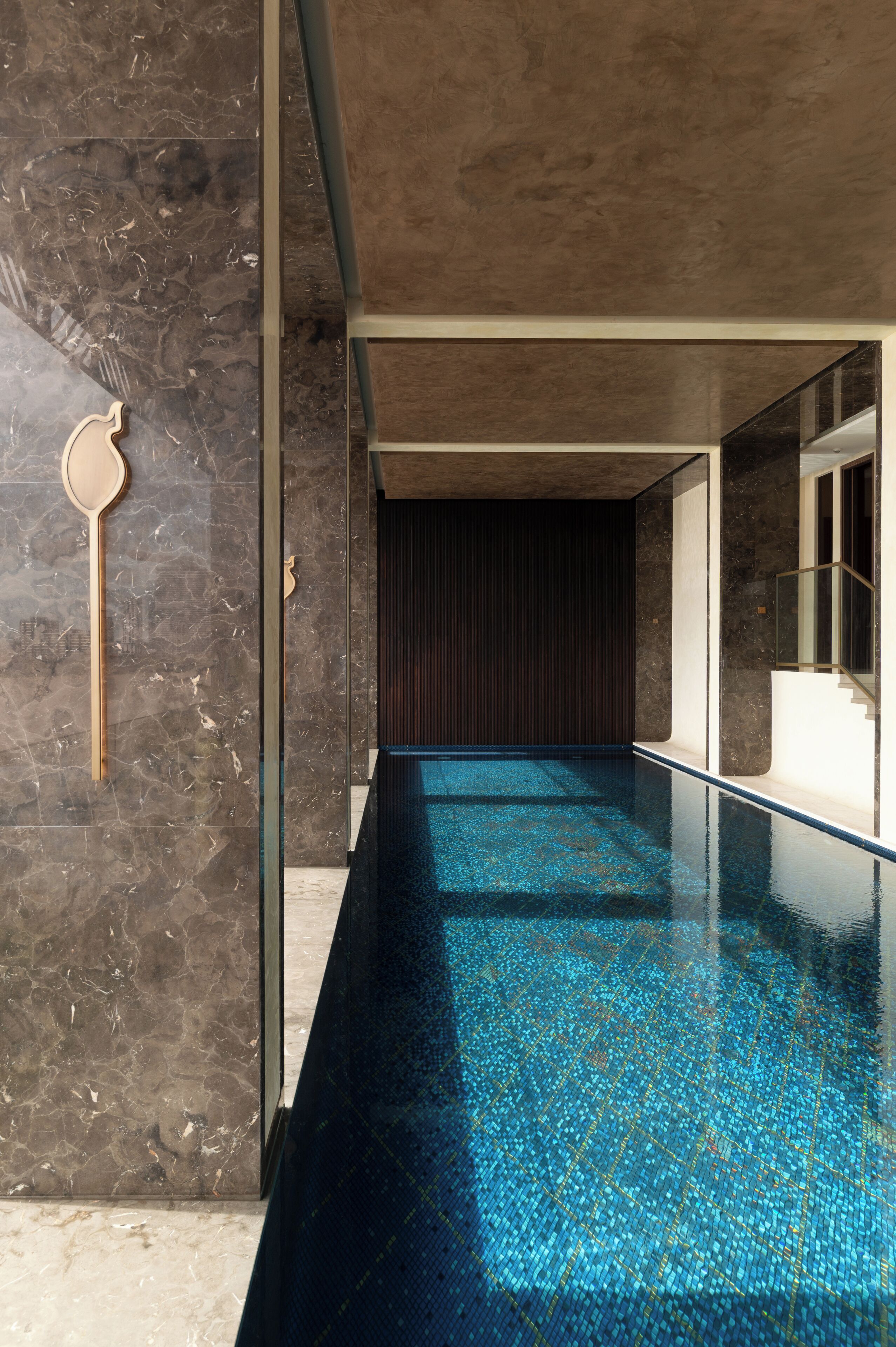 A modern indoor swimming pool with a dark floor and clear blue water, surrounded by marble walls and a glass barrier.