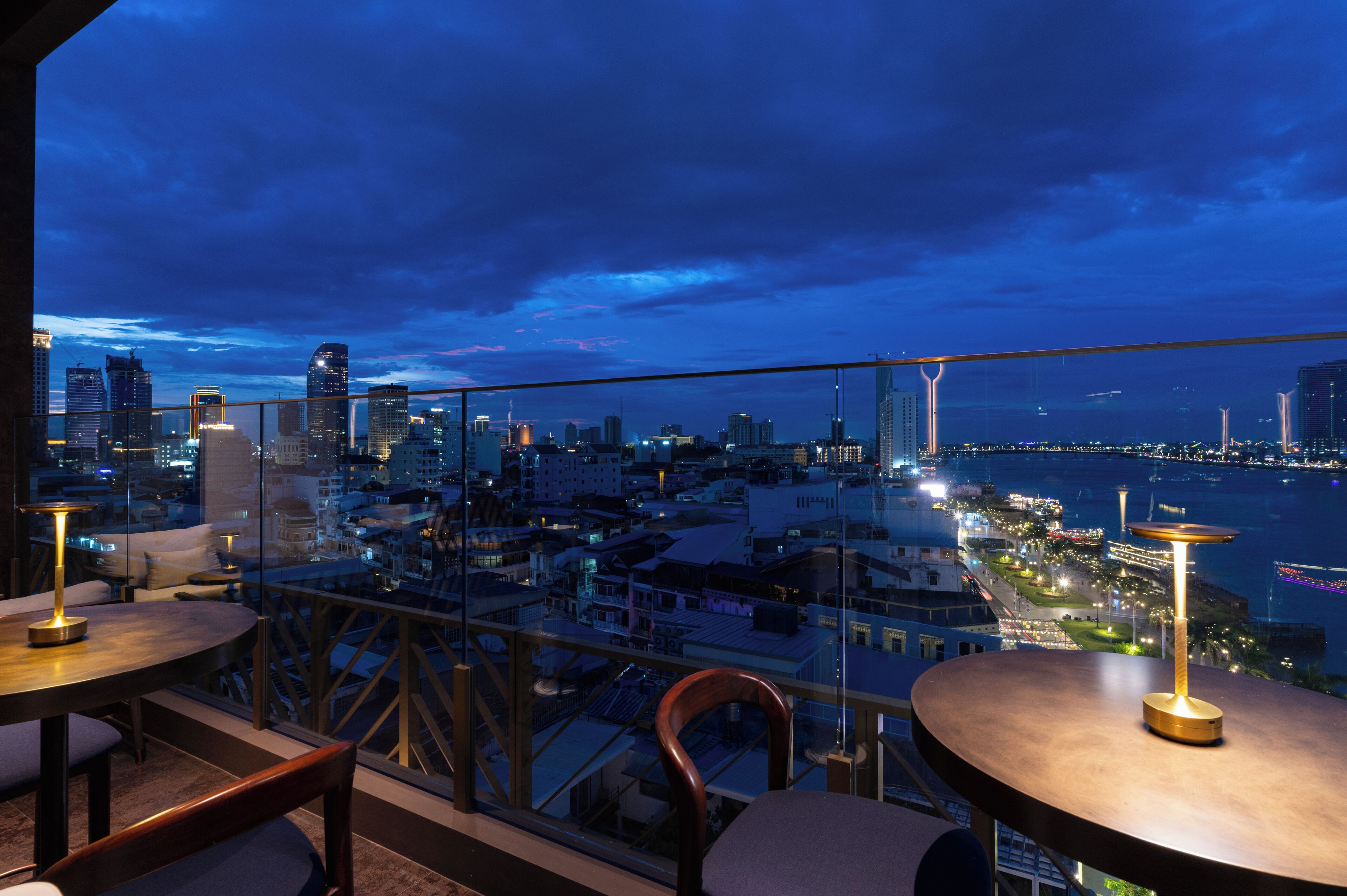 A rooftop terrace with tables and chairs, overlooking a cityscape with buildings and a river.