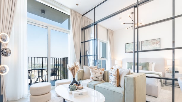 Ein modernes Wohnzimmer mit Sofa, Hocker und Couchtisch. Durch große Fenster ist ein Balkon mit Gartenmöbeln zu sehen.